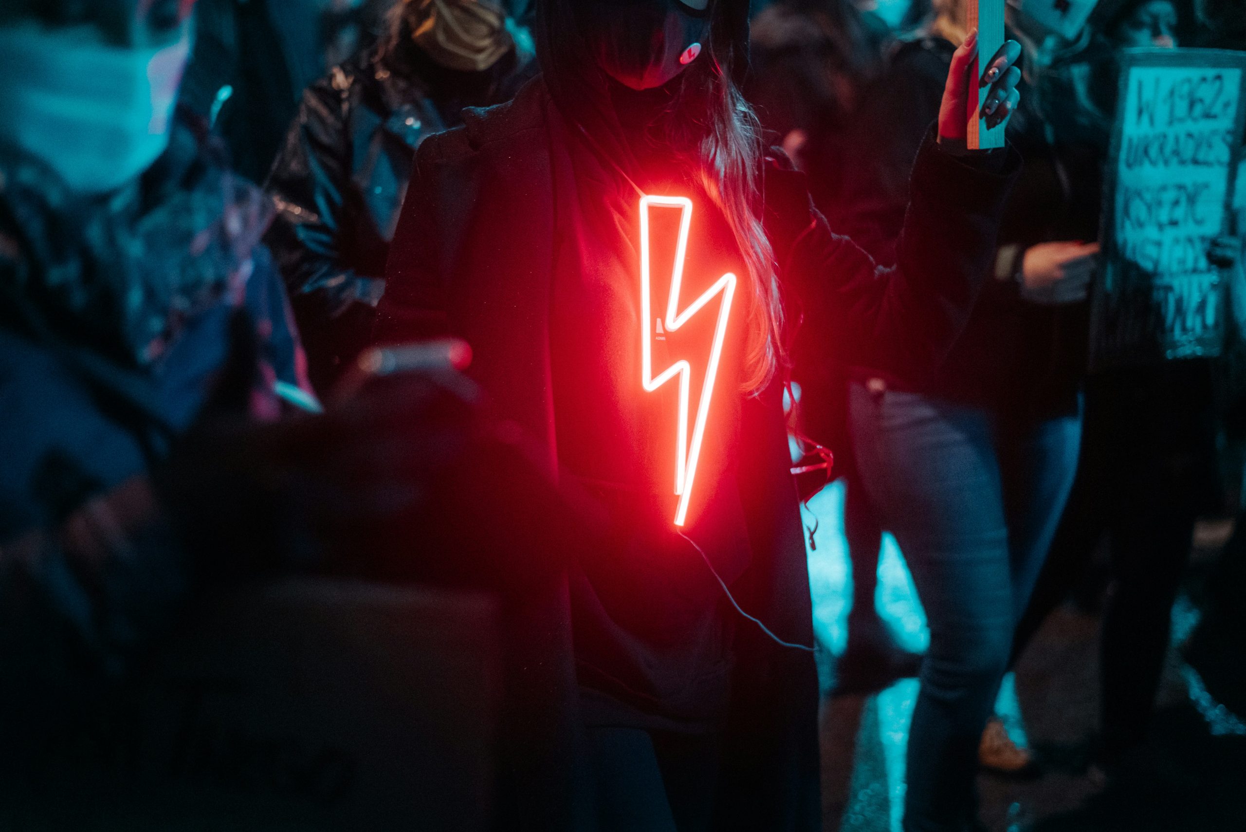 Close cropped image of people protesting. One protestor is wearing a neon sign that is a lightning bolt.